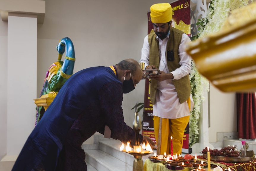 A man in wearing  a purple long shirt bends in front of row of lights while a man wearing traditional Indian clothes looks on.