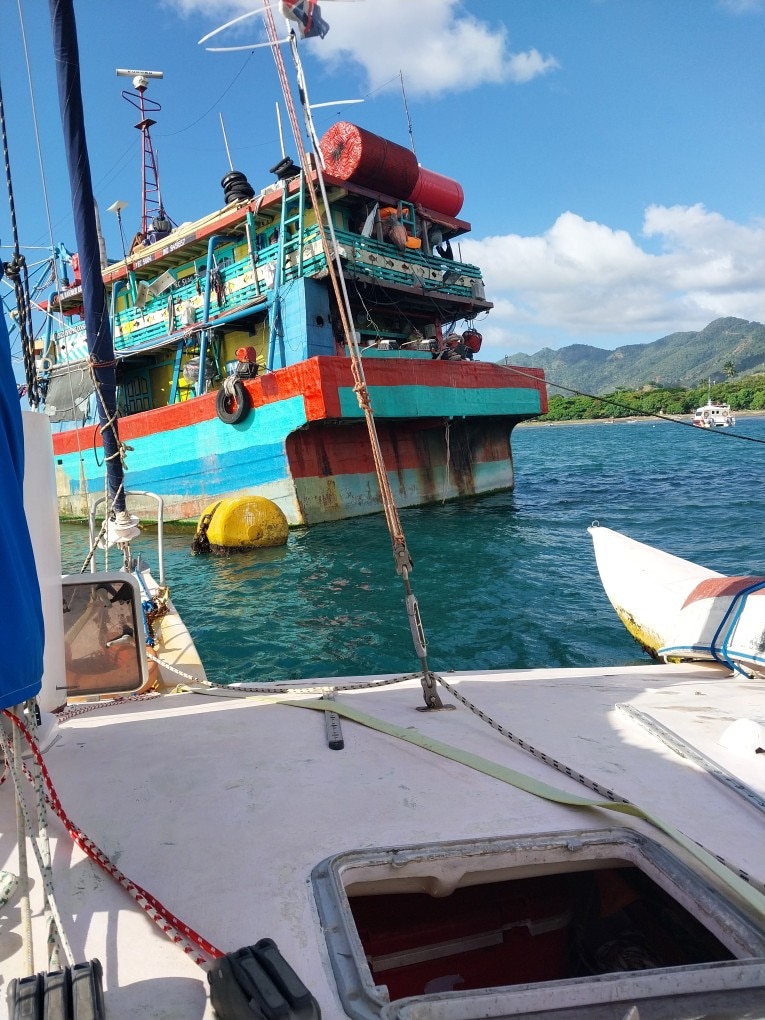 The view from a trimaran looking at a big boat that is blue, red, off of a coast with mountains in the background.