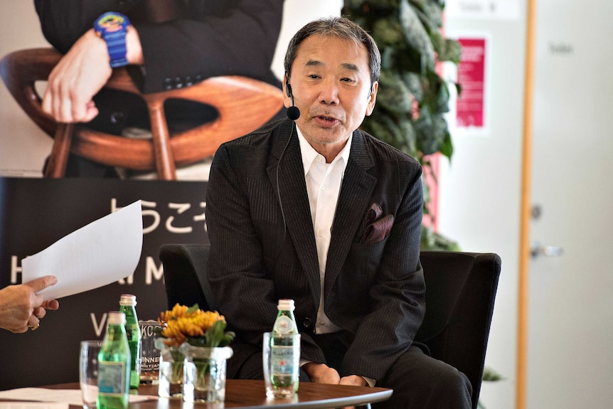Haruki Murakami sits at a table wearing a an earpiece microphone.