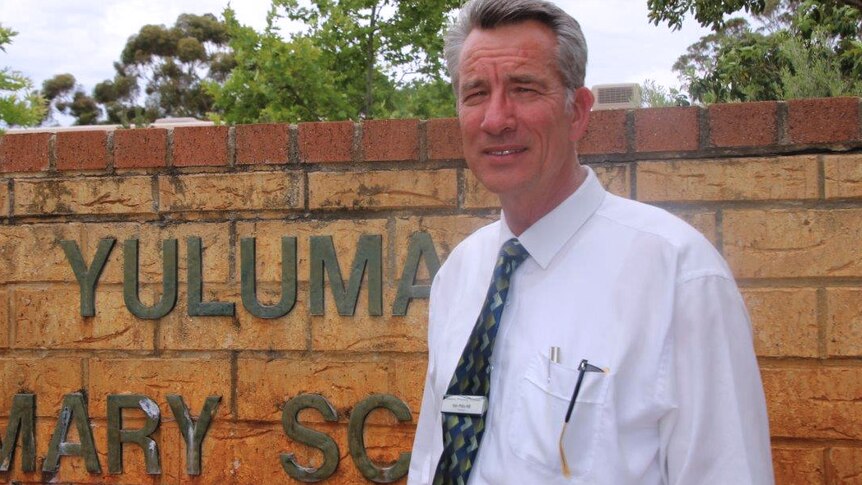Kim Pitts-Hill poses for a photo standing in front of a brick wall with Yuluma Primary School written on it.