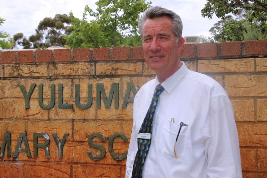Kim Pitts-Hill poses for a photo standing in front of a brick wall with Yuluma Primary School written on it.