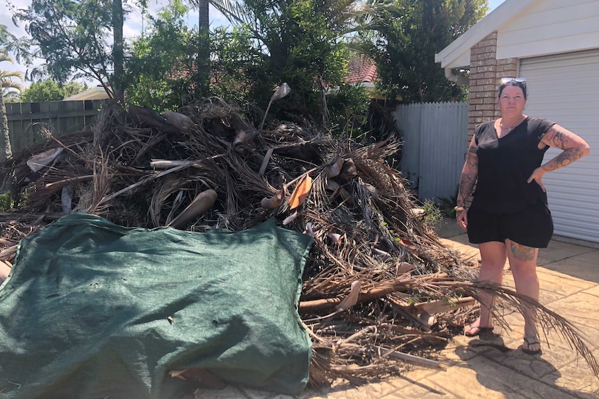 Jade Smith stands next to a large pile of palm fronds and tree branches on the driveway of her home.
