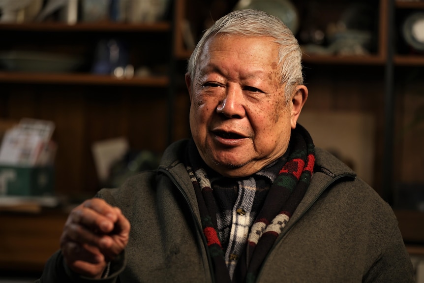 A man with short grey hair sits in a warm lit room during an interview