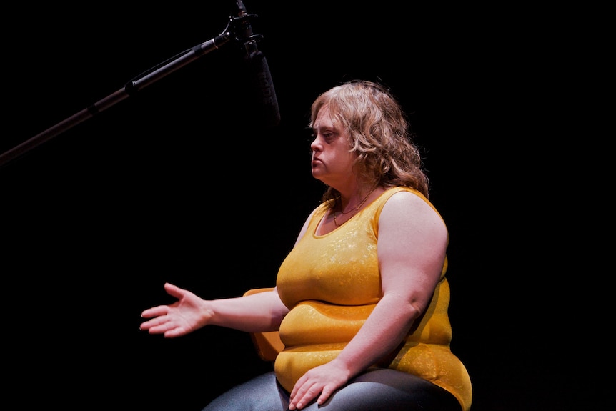 A woman on stage, microphone aimed at her, she reaches out to the audience in Back to Back Theatre's production of Food Court
