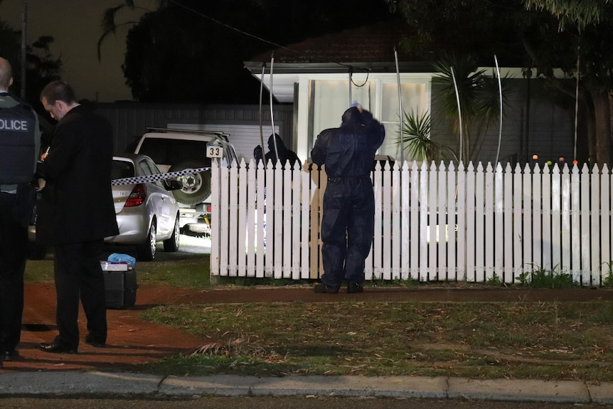A police forensic officer stands next to a white picket fence outside a house at night, with officers in the driveway nearby.