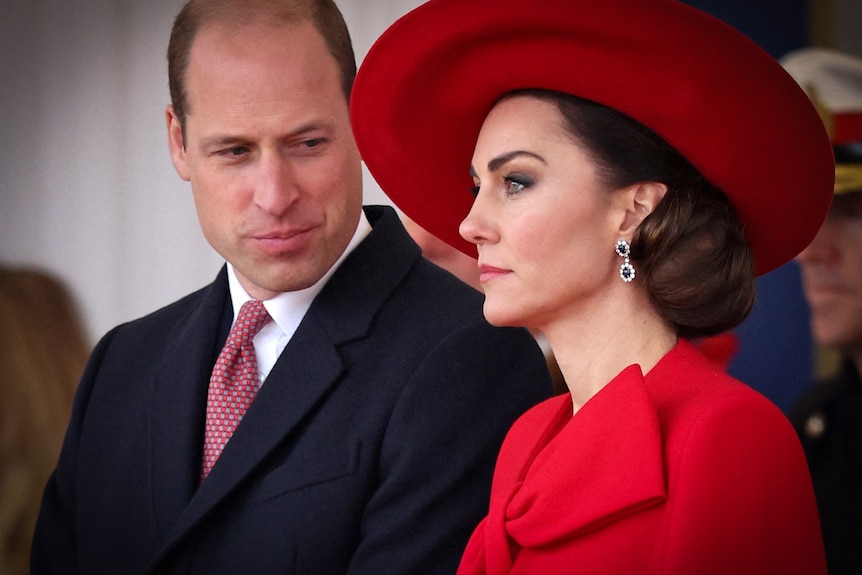 A close up of Prince William smiling down at his wife Catherine, who is wearing a red dress and hat.
