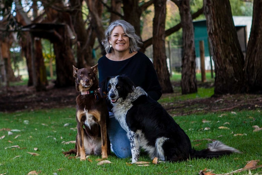 A middle-aged woman with a big smile kneels on the ground with her two dogs.