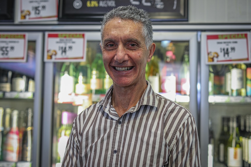 A man smiles in front of a fridge of alcohol.