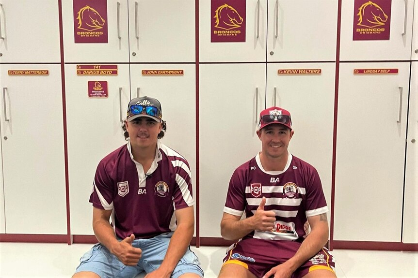 Two young men sit on a bench wearing brown shirts in front of white lockers with Broncos rugby league stickers on them