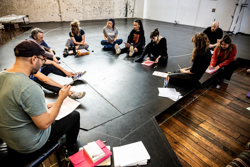 A group of people wearing casual clothes and sitting in a circle in a rehearsal space, holding scripts in their hands