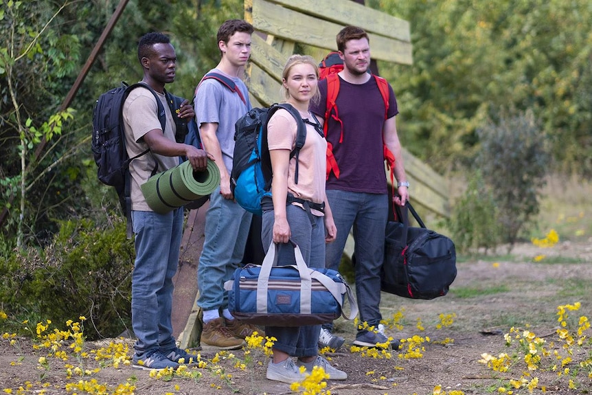 The four actors stand in the wilderness carrying luggage in the classic European backpacker style.