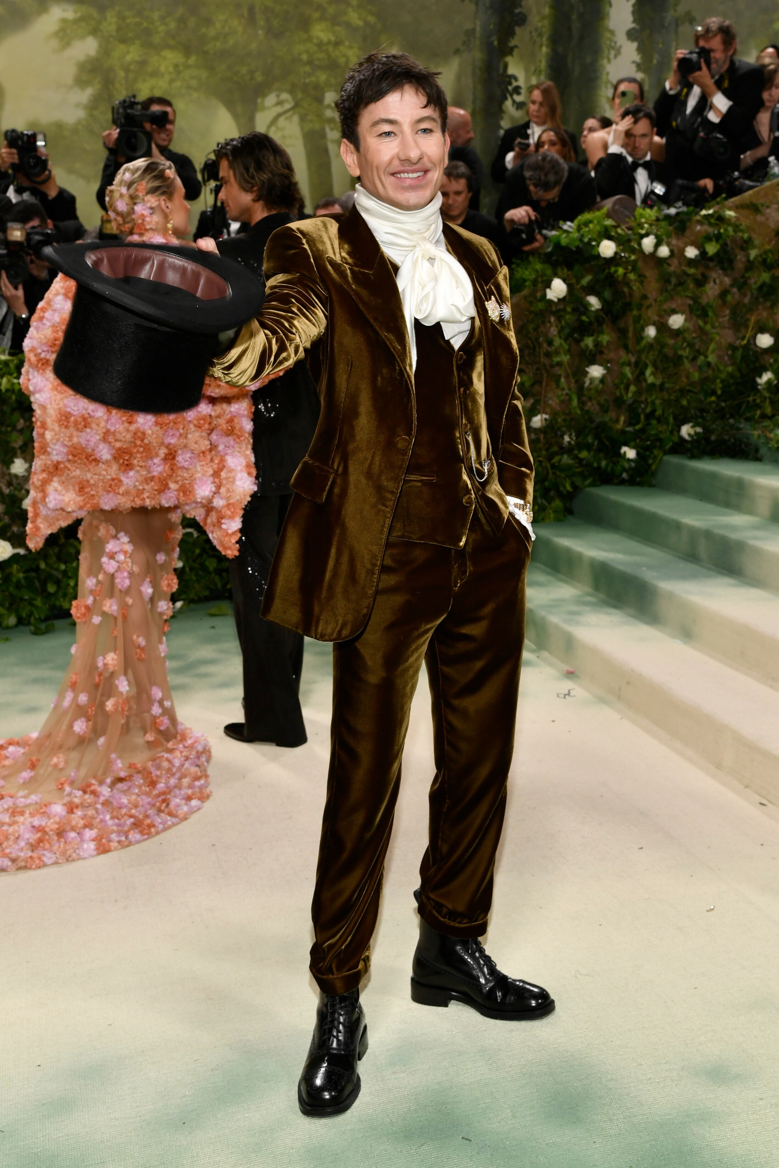 Barry Keoghan wearing a tan velvet suit with a white, ruffly scarf wrapped around his neck, holding a top hat.