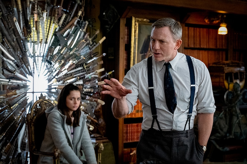 Daniel Craig stands with one hand raised in ornate room with Ana de Armas who sits on chair near collection of knives and axes.