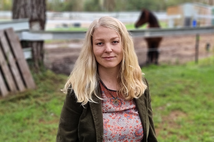 A young woman with long blonde hair looks into the camera with a serious expression. A horse is in the background.