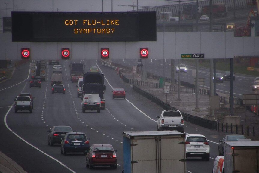 Grey skies above traffic on a freeway with a gantry sign saying "Got flu-like symptoms?"