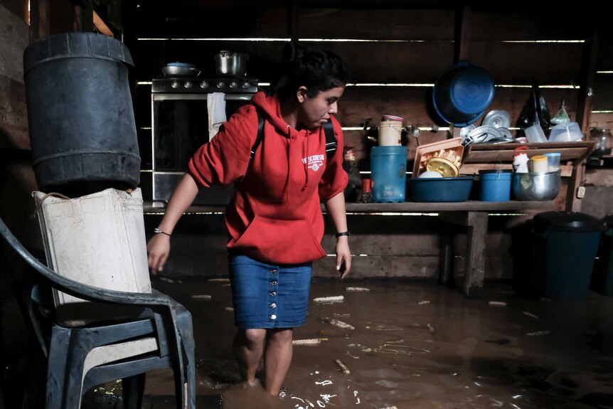 A woman in a red jumper wades through ankle-high water in a wooden house.