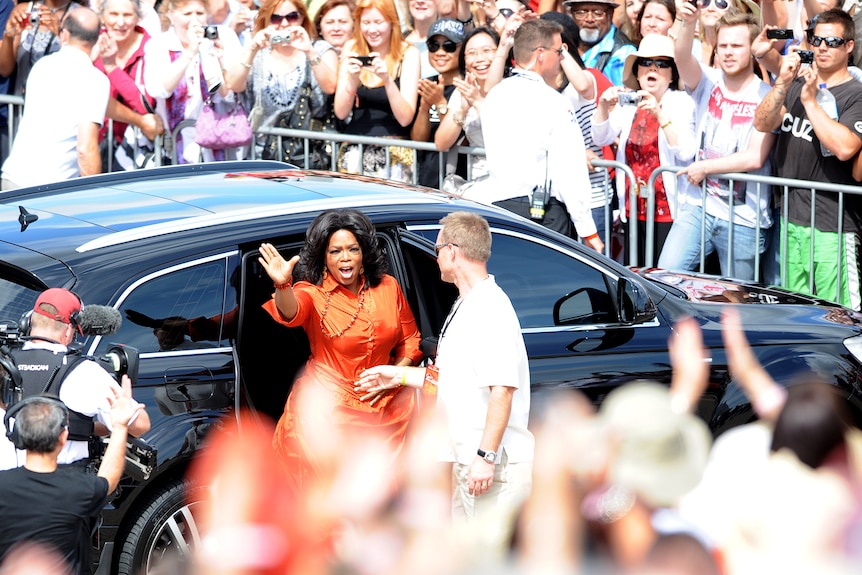 Oprah Winfrey dressed in orange gets out of a black car surrounded by fans