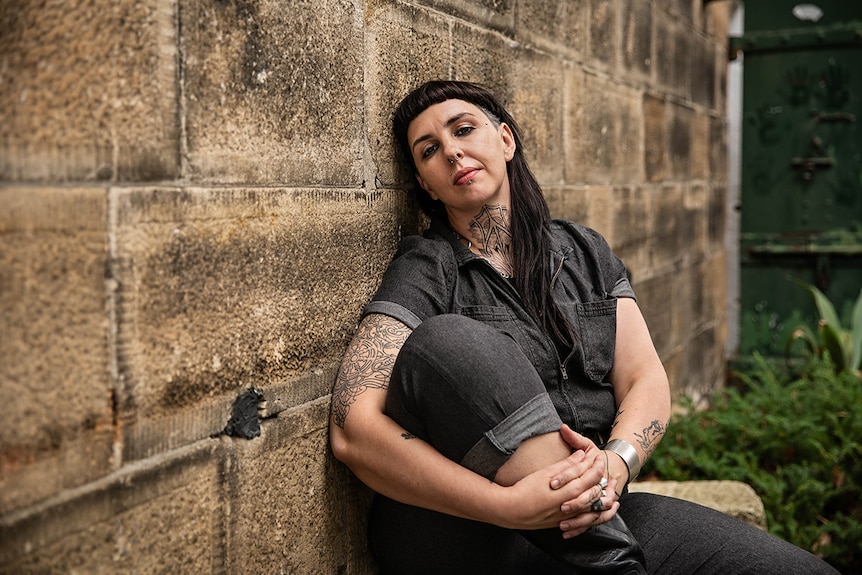 Colour photo of artist S.J Norman sitting against sandstone wall at the National Art School in Sydney.