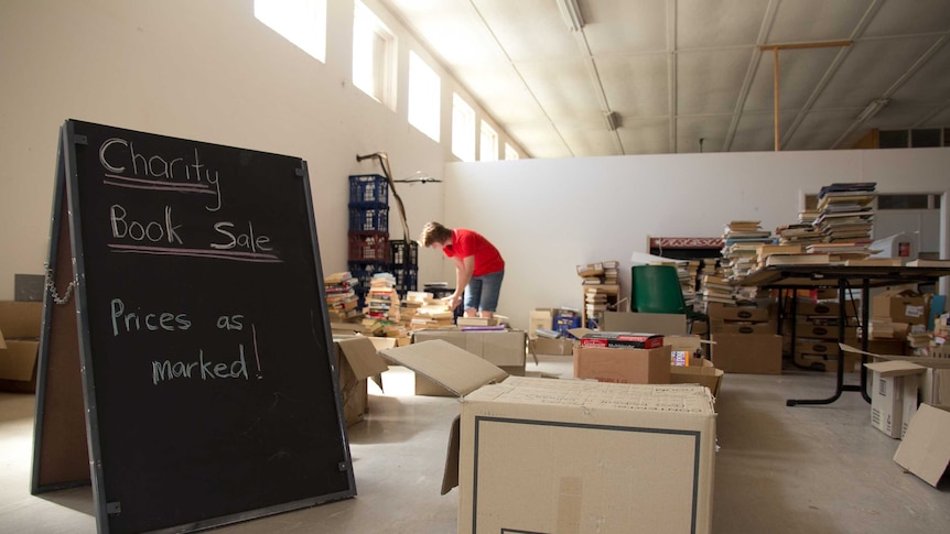 A chalkboard with the words Charity Book sale in the foreground with boxes of books in the background a woman looking at them