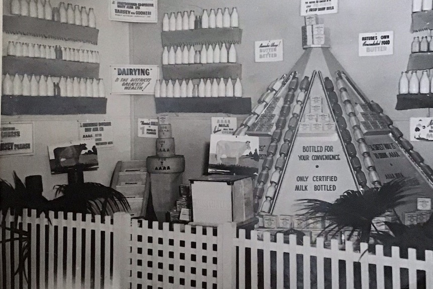 glass milk bottles stacked in a triangular tower and bottles line the wall, behind a small picket fence