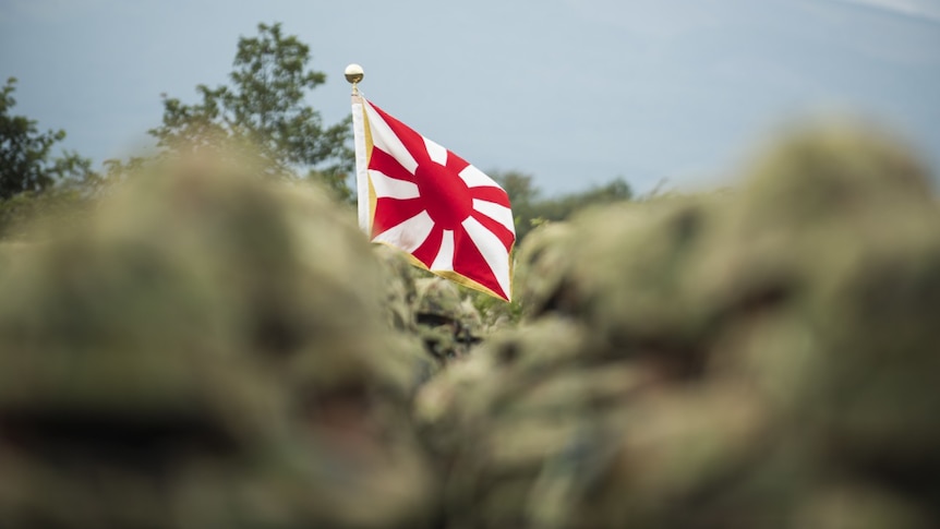A flag of the Japanese Self-Defence forces appears through a blurred crowd of service personnel.