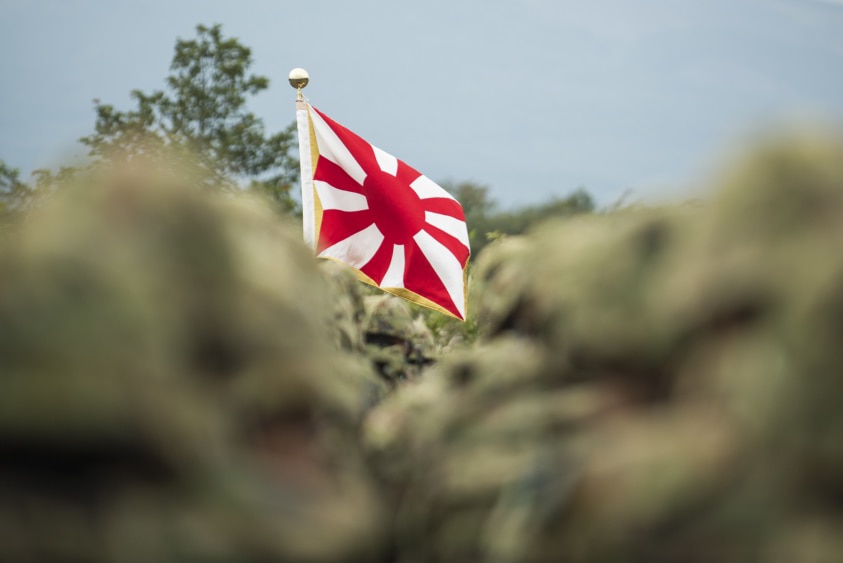 A flag of the Japanese Self-Defence forces appears through a blurred crowd of service personnel.