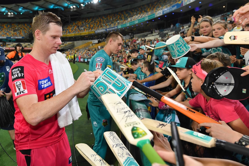 A star cricketer with a towel flung over his shoulder signs autographs for fans after a BBL game.