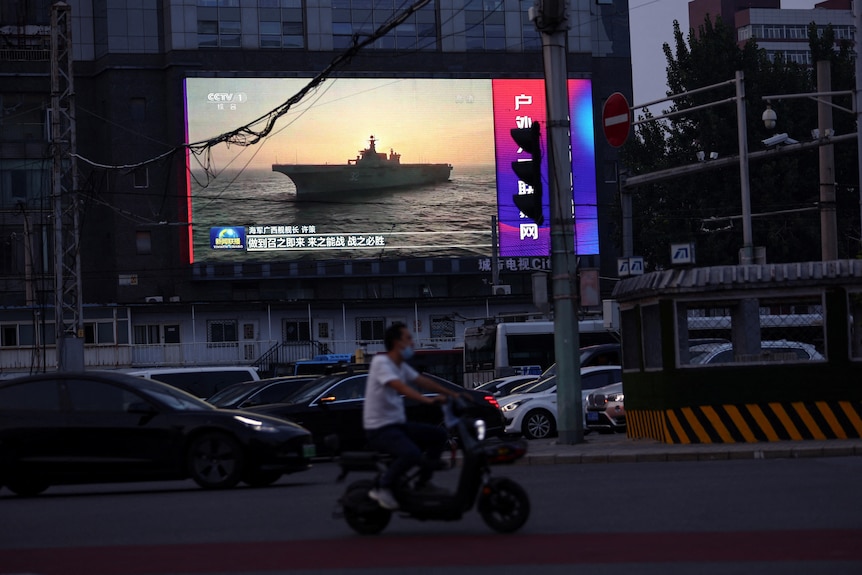 Mężczyzna jedzie na motocyklu przed ekranem pokazującym materiał z Chińskiej Armii Ludowo-Wyzwoleńczej