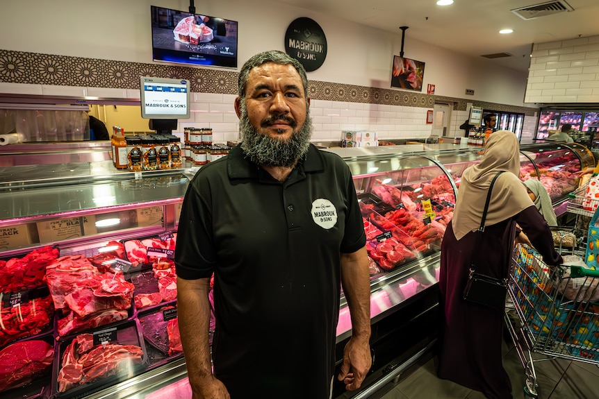 A bearded man in a dark polo shirt stands in a butcher's shop.