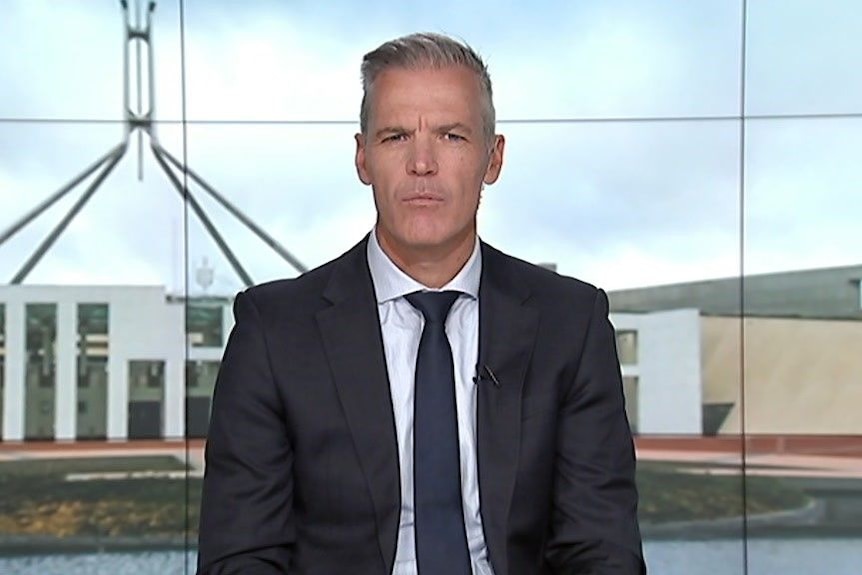 A grey-haired man in a dark suit sits in a television studio with a parliament house backdrop.
