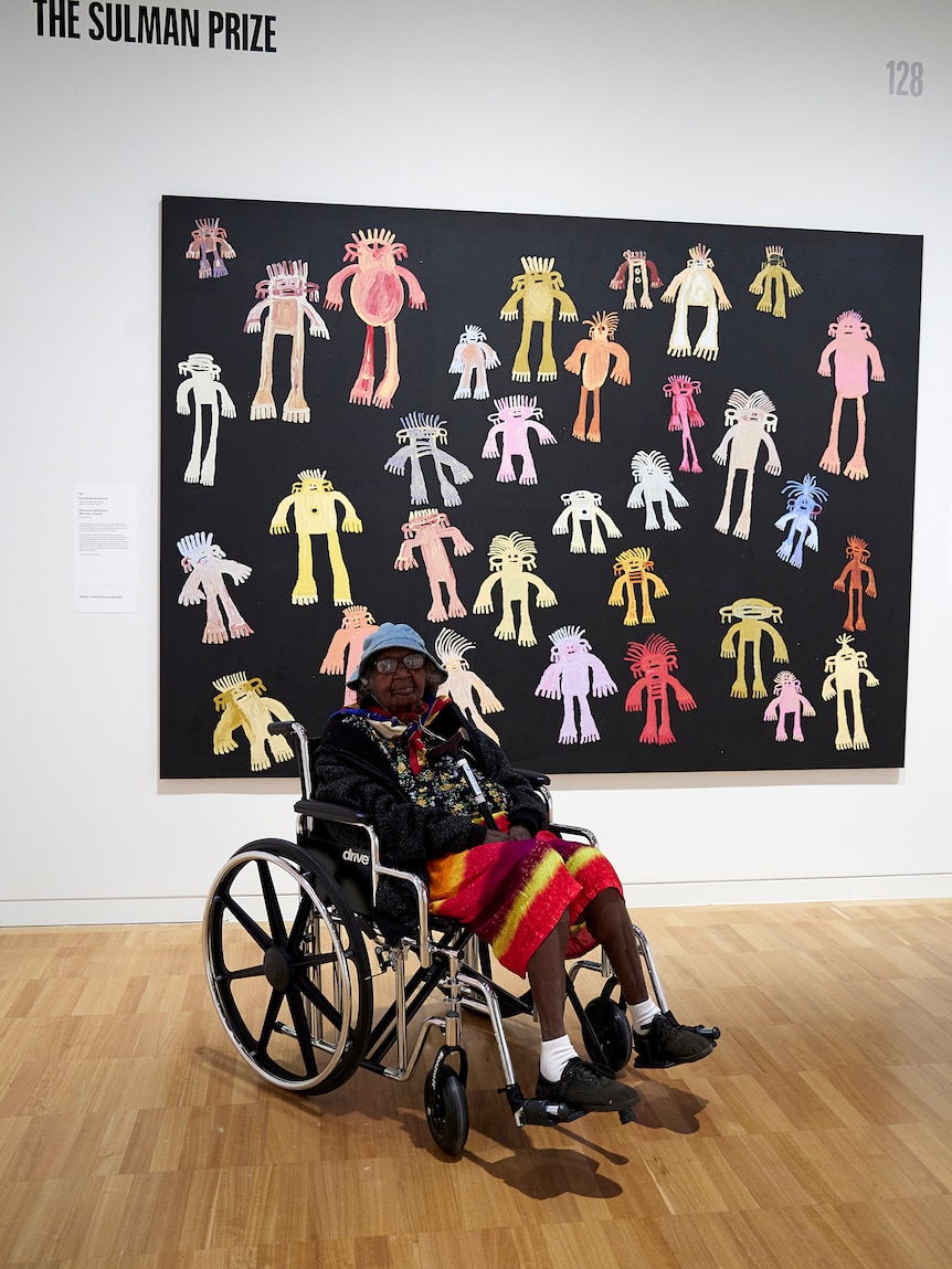 An Indigenous Australian woman in bright clothing sits in a wheelchair in front of a large artwork.