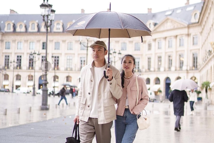 A man wearing a white shirt and jacket holds an umbrella while walking next to a woman wearing a pink jacket and jeans.