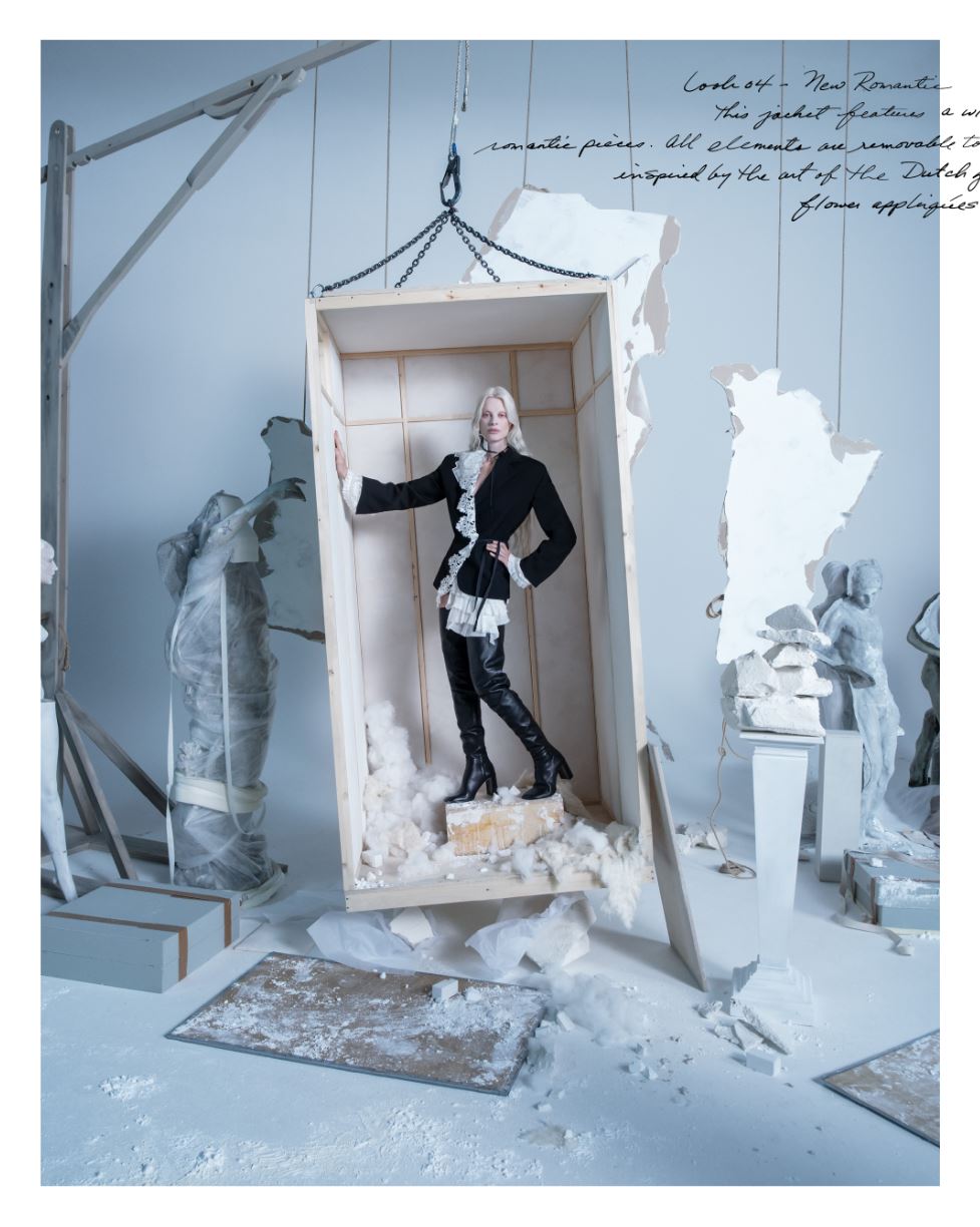 A woman in a black suit stands in a wooden packing crate, surrounded by plaster rubble and statues. 