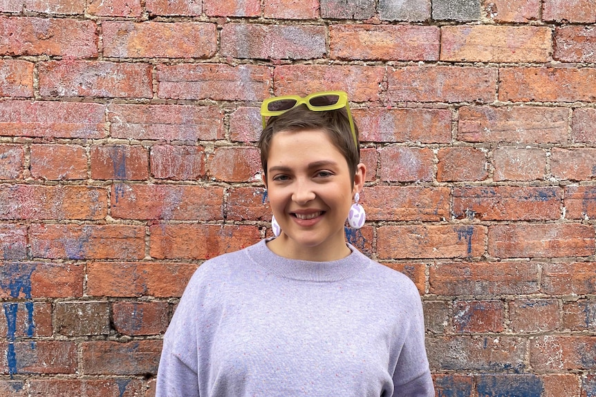 A young woman with short cropped hair and pastel purple knit jumper in front of a red brick exterior wall. 