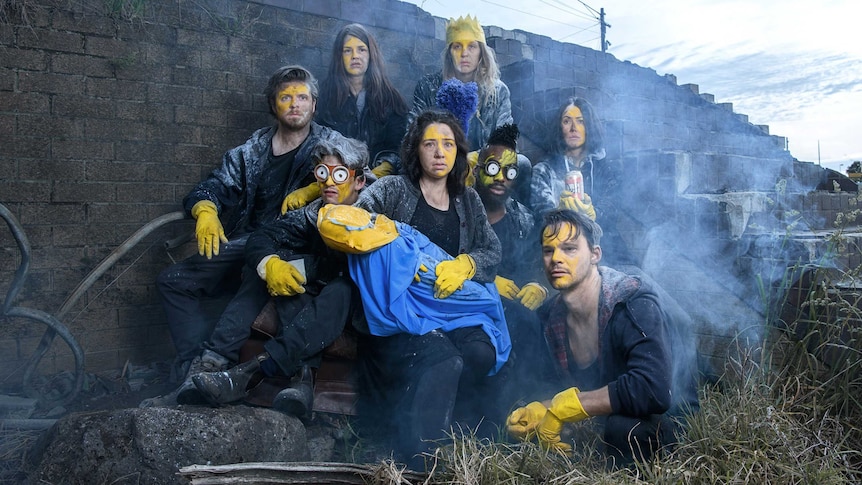 Eight people wearing yellow gloves and yellow face paint huddle by a dilapidated brick staircase outside.