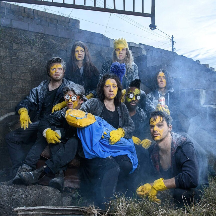 Eight people wearing yellow gloves and yellow face paint huddle by a dilapidated brick staircase outside.