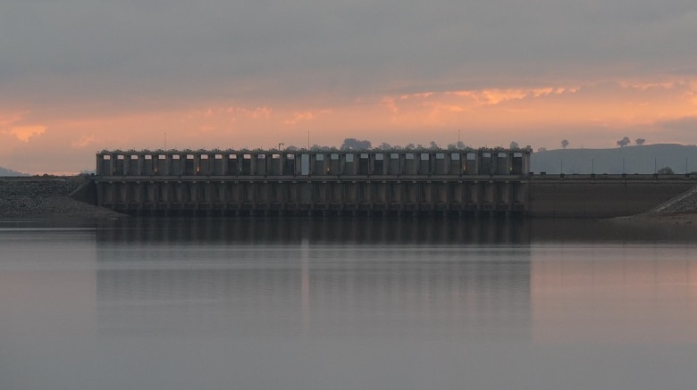 A large body of still water sits in the foreground. The large concrete walls of hum dam are in the mid line, seen from behind.