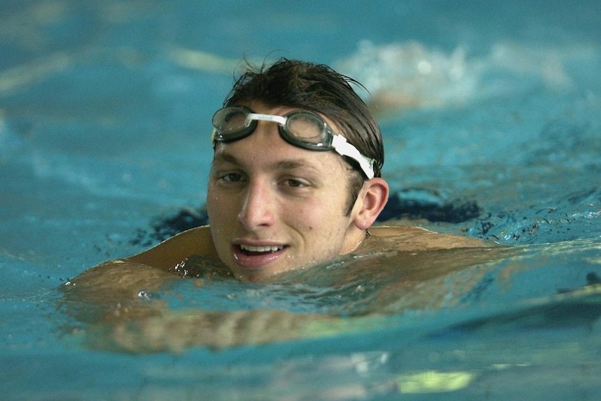 Ian Thorpe swims with his head out the water and goggles on his forehead, arms out in front, and smiles