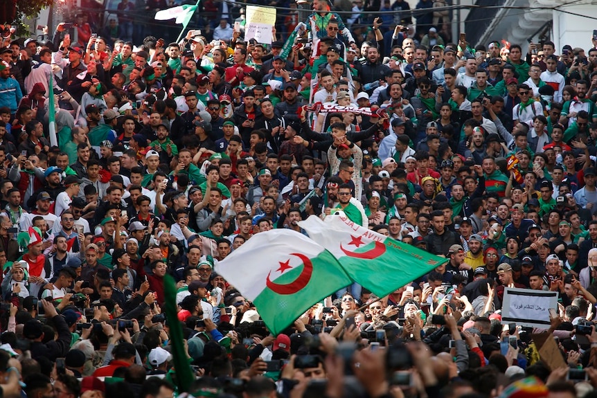 A large crowd of protesters look towards the same point as some wave the green, white and red Algerian flag.