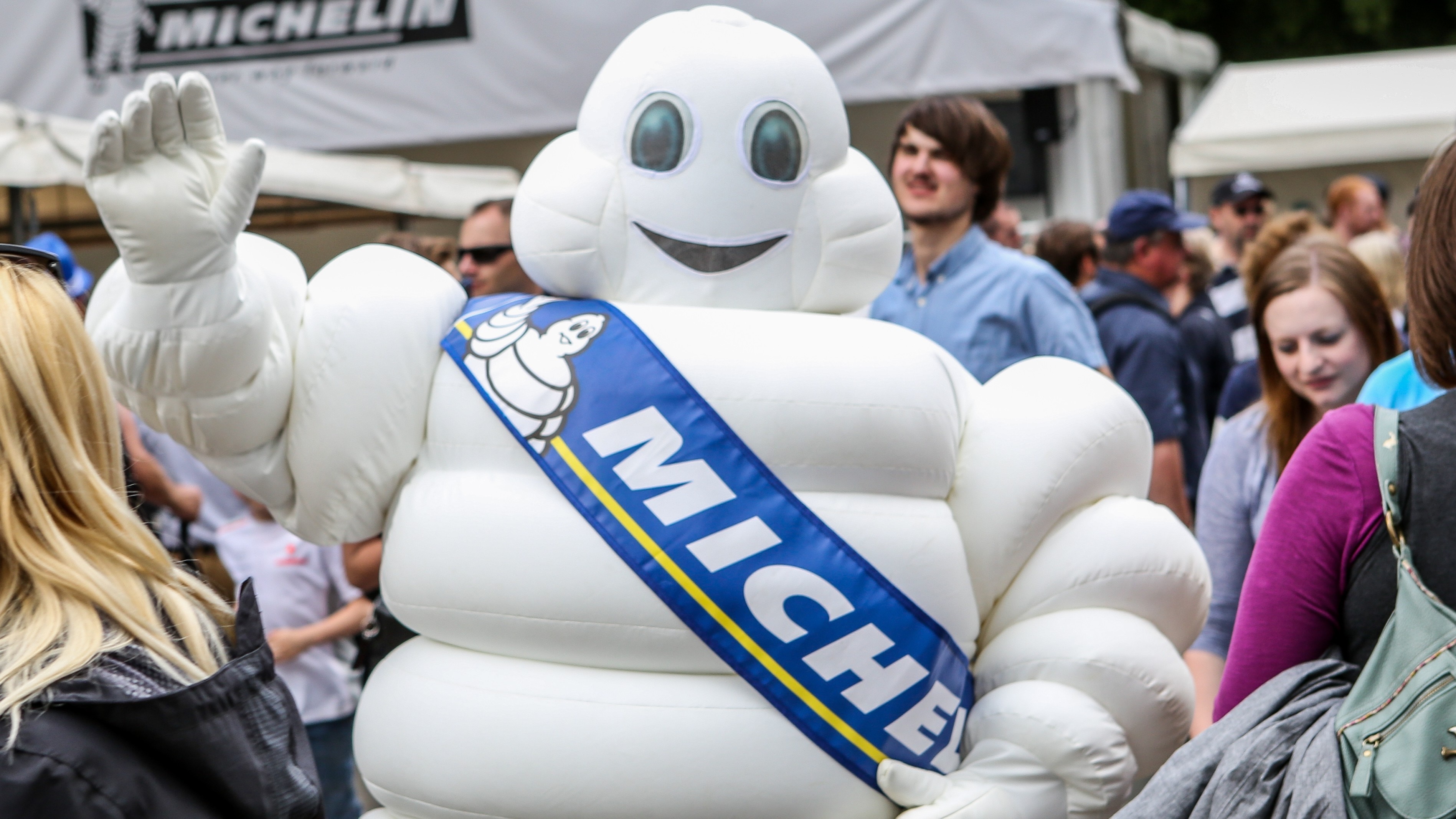 The Michelin Man, a white puffy mascot for the Michelin tyre company, wearing a blue sash and waving at the camera
