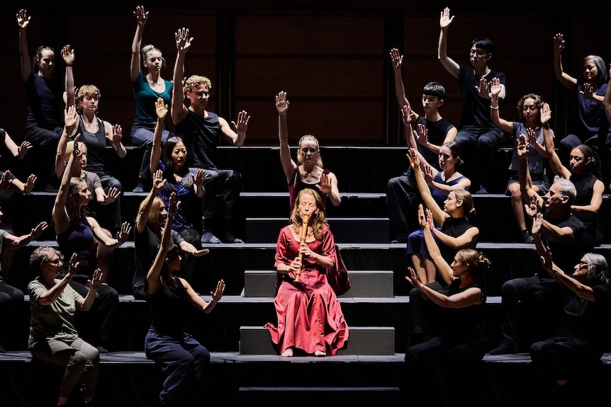 A woman in a red dress plays the recorder on stage, surrounded by people in black outfits with their arms raised.