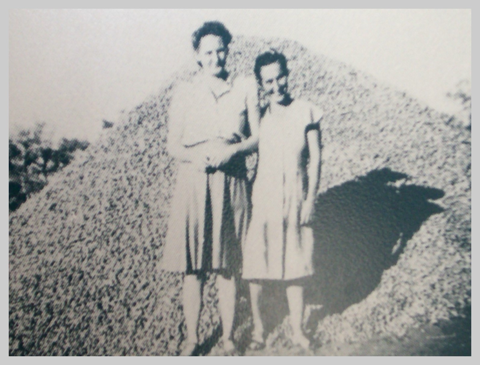 Two women, on taller than the other, stand in front of a pile of peanuts almost as tall as them, in black and white