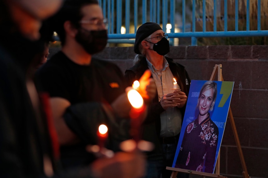 Men in black face masks attend an evening vigil for Halyna Hutchins, holding bright orange candles beside a picture of her 