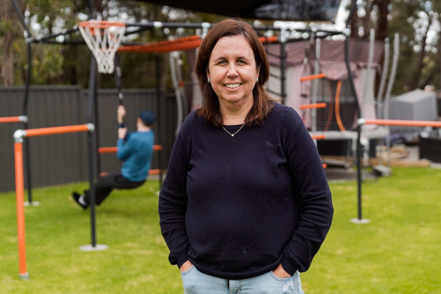 A middle-aged woman with brown hair and a blue jumper stands in a backyard smiling in front of a boy playing on a swing.