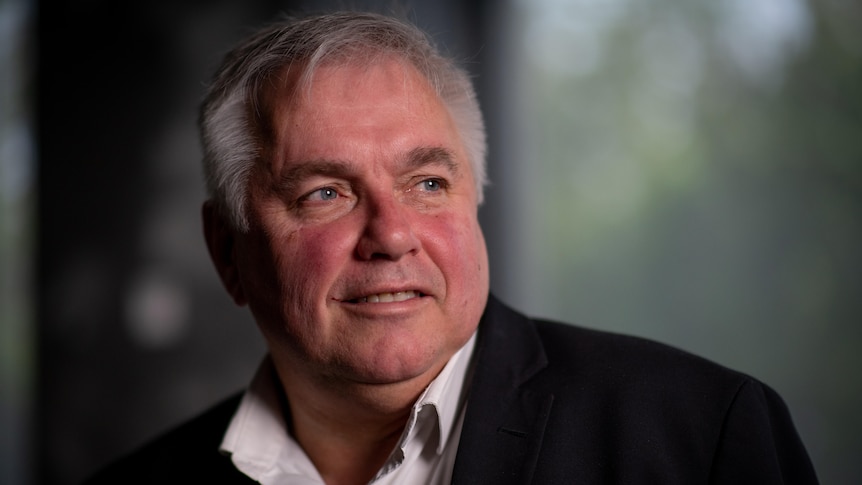 A man with grey hair wearing a black jacket and white open-necked shirt