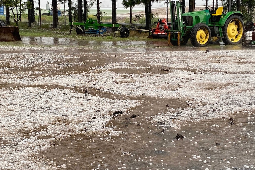 Hail covers the ground near a tractor and other farm machinery.