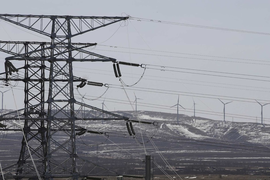 You view large transmission power lines with a snowy hill of wind turbines on the horizon on an overcast day.