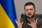 Volodymyr Zelenksyy speaks in front of a Ukrainian flag.
