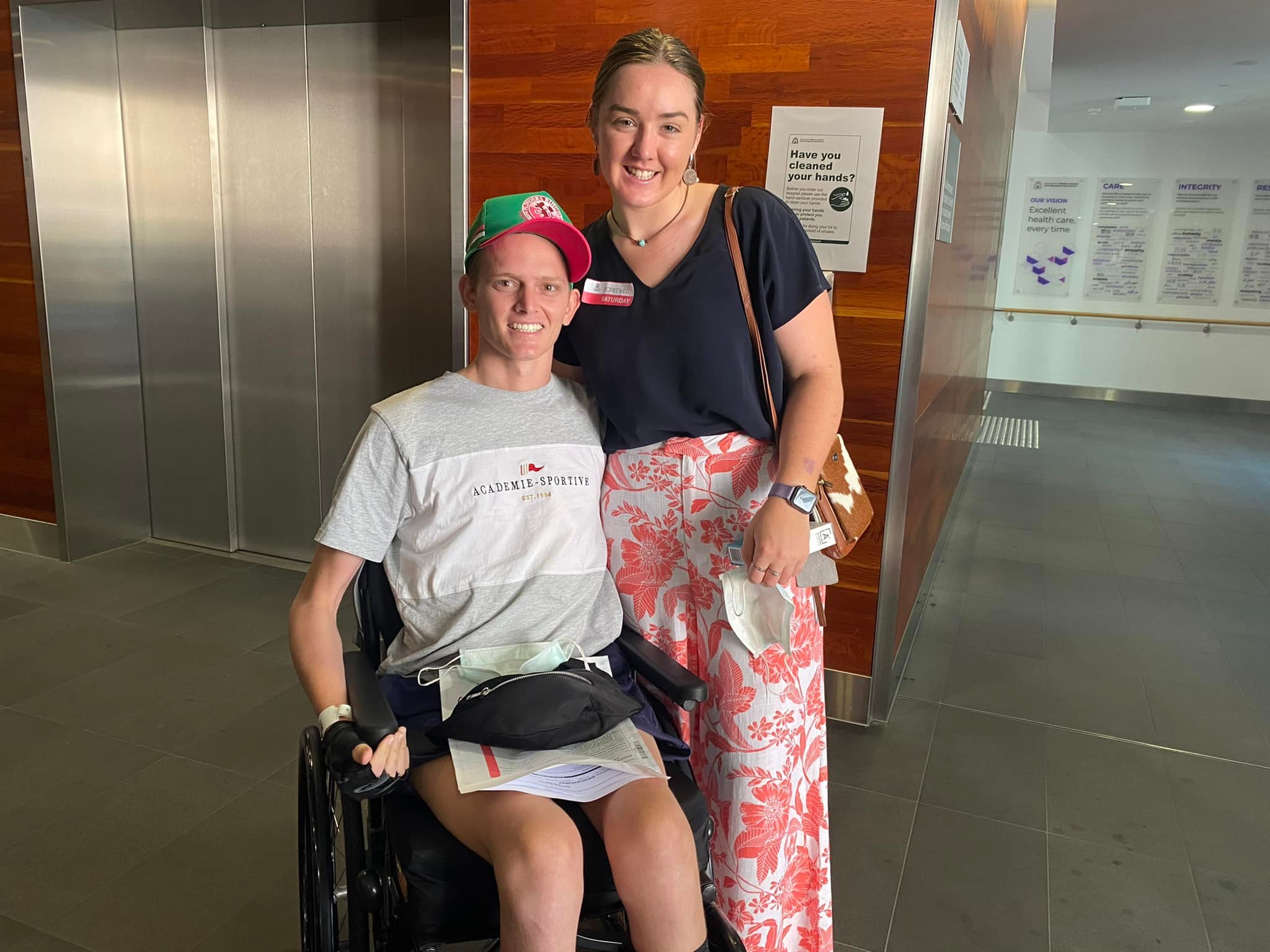 A smiling young couple, she stands beside him as he sits in a wheelchair, with thin arms.