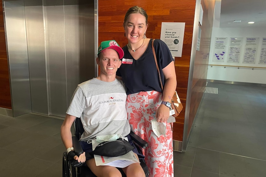 A smiling young couple, she stands beside him as he sits in a wheelchair, with thin arms.